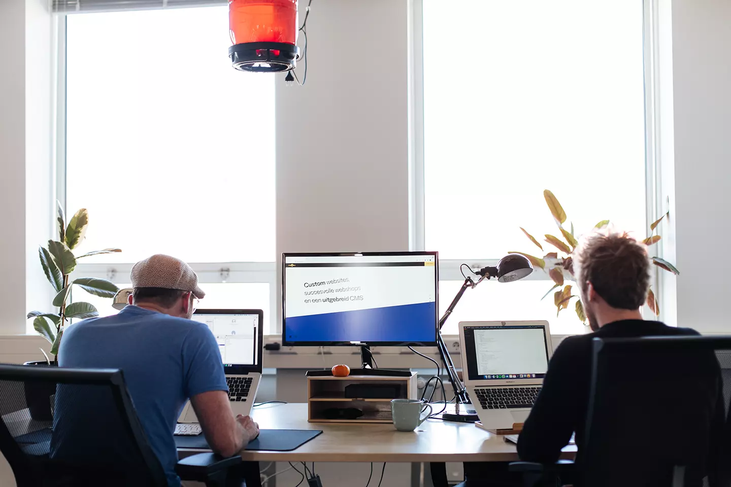 Foto van twee jonge mannen aan het werk achter hun laptop. Groot computer scherm tussen hen in